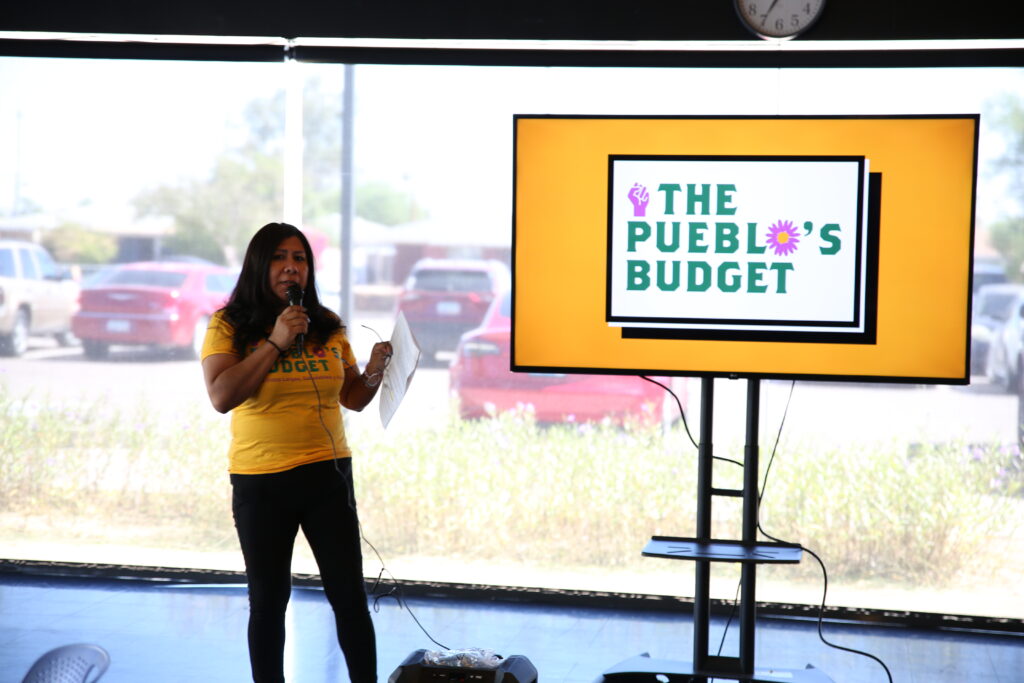 A woman speaking into a microphone, standing next to a TV screen that says "The Pueblo's Budget"